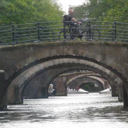 En person cyklar på en tegelbågsbro över en kanal med flera bågbroar och träd i bakgrunden.
