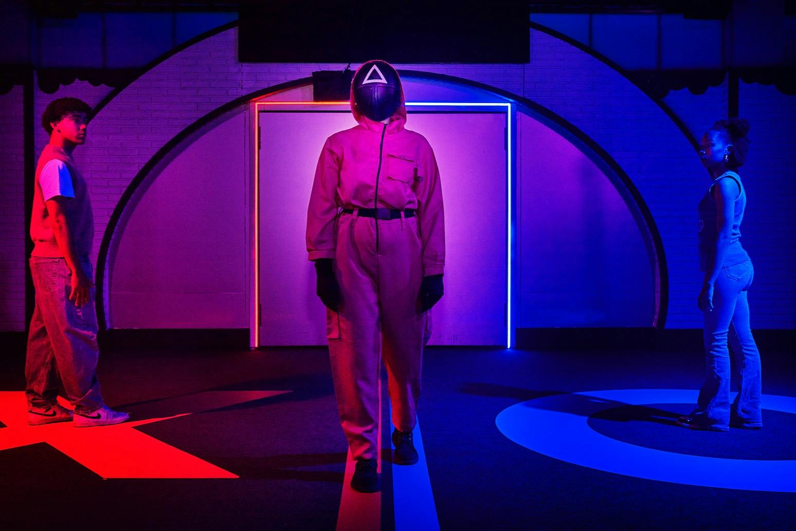 一个穿着红色连身裤，戴着黑色头盔的人站在灯火通明的房间里，地板上有红色和蓝色的灯光和几何形状。