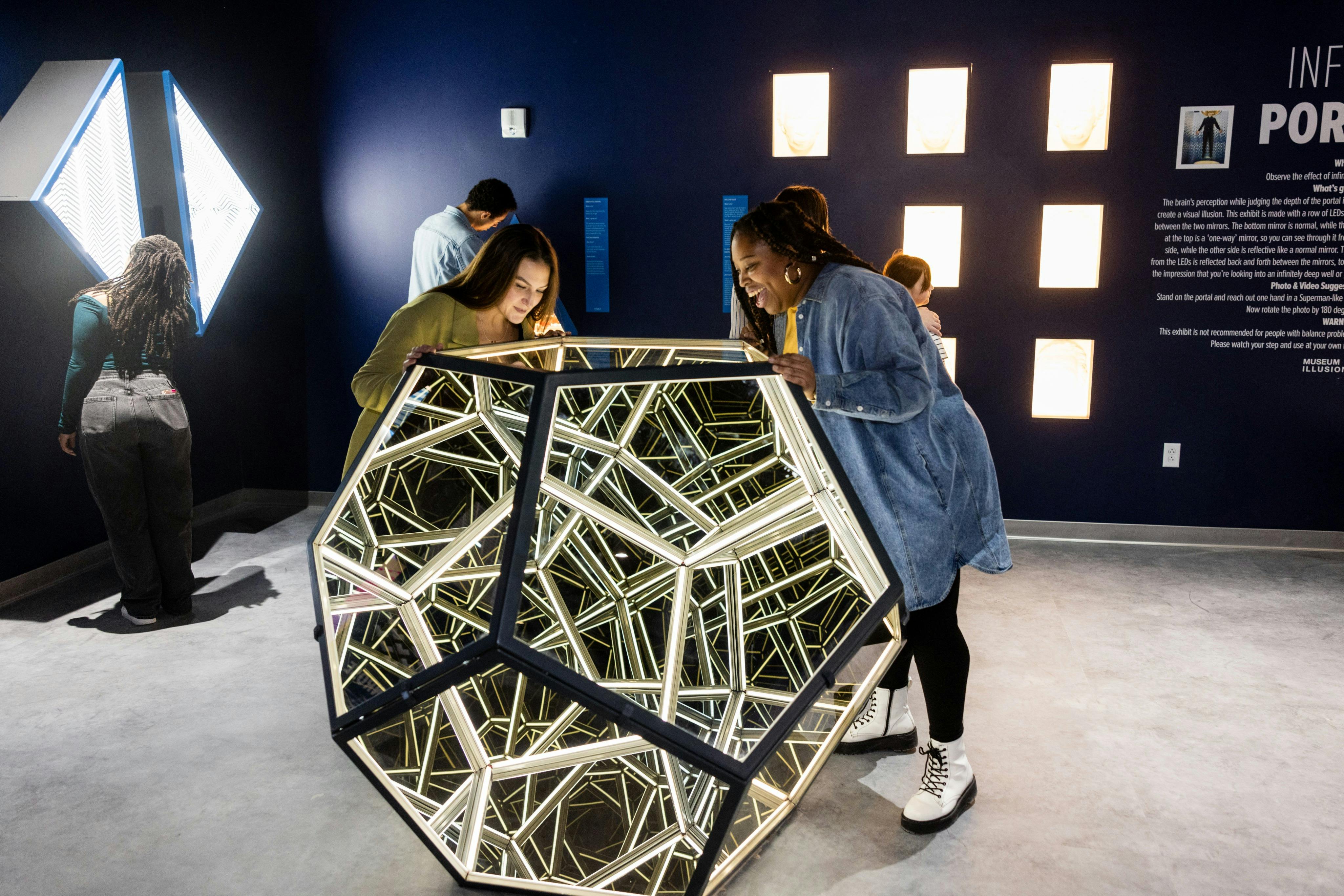 Mensen kijken in een geometrisch gespiegeld beeld in een moderne tentoonstelling met donkerblauwe muren en verlichte displays.