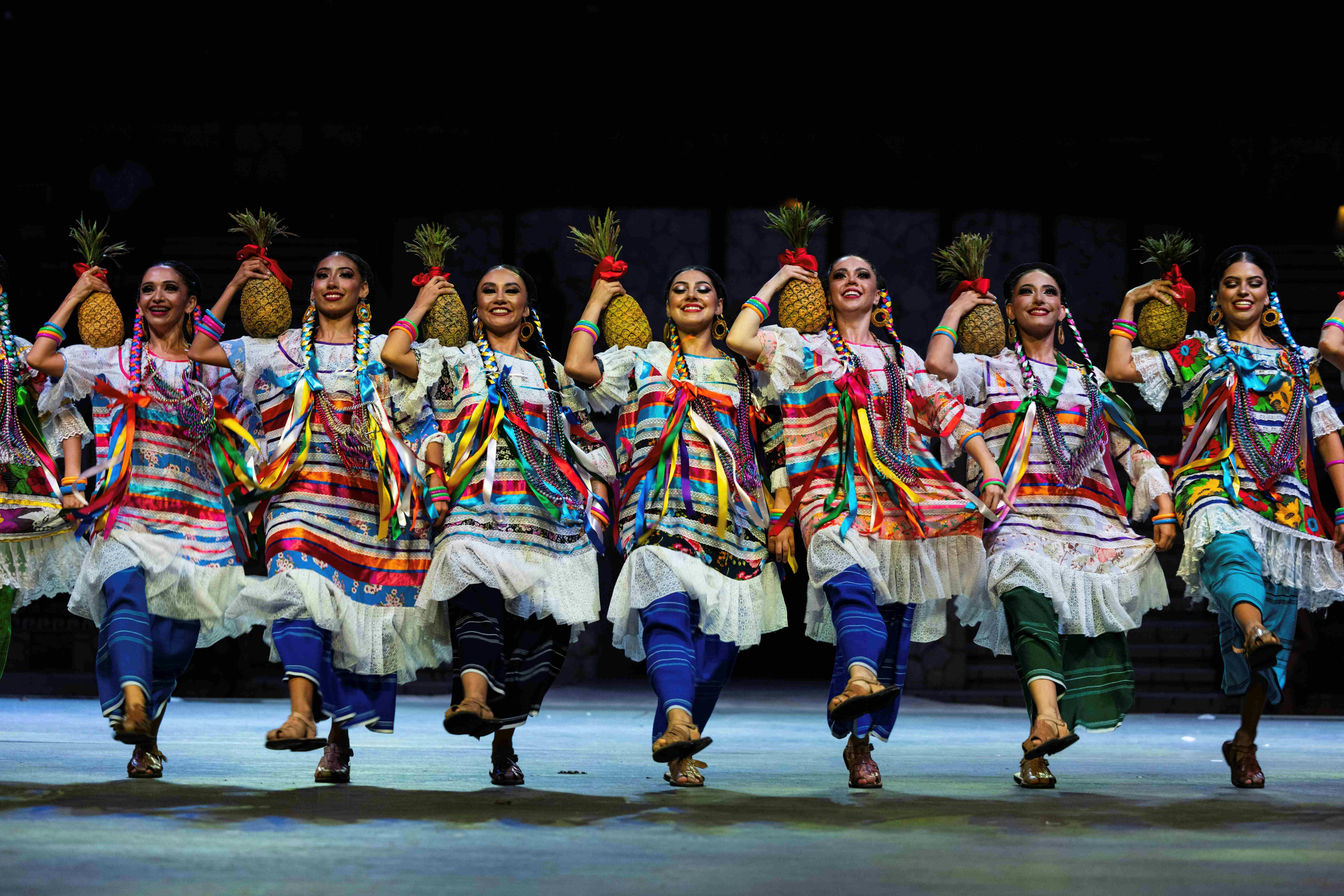 A group of dancers in colorful traditional attire, holding pineapples, performing on a stage with coordinated movements.