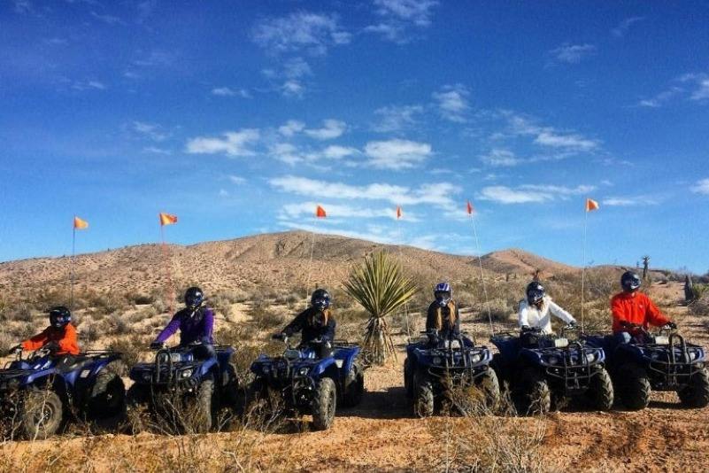 Four people on ATVs in a desert landscape with a mountain in the background under a clear blue sky.