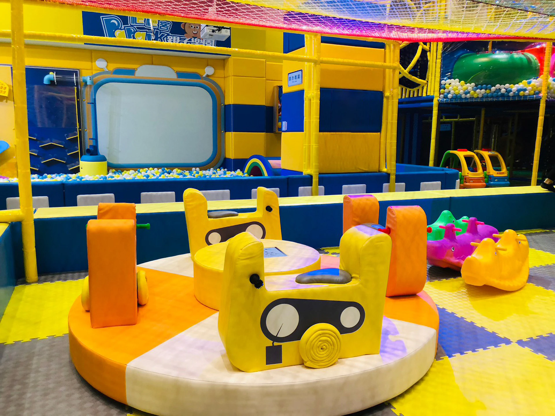 Brightly colored indoor children's play area with padded seating, a ball pit, and playful structures under a netted ceiling.