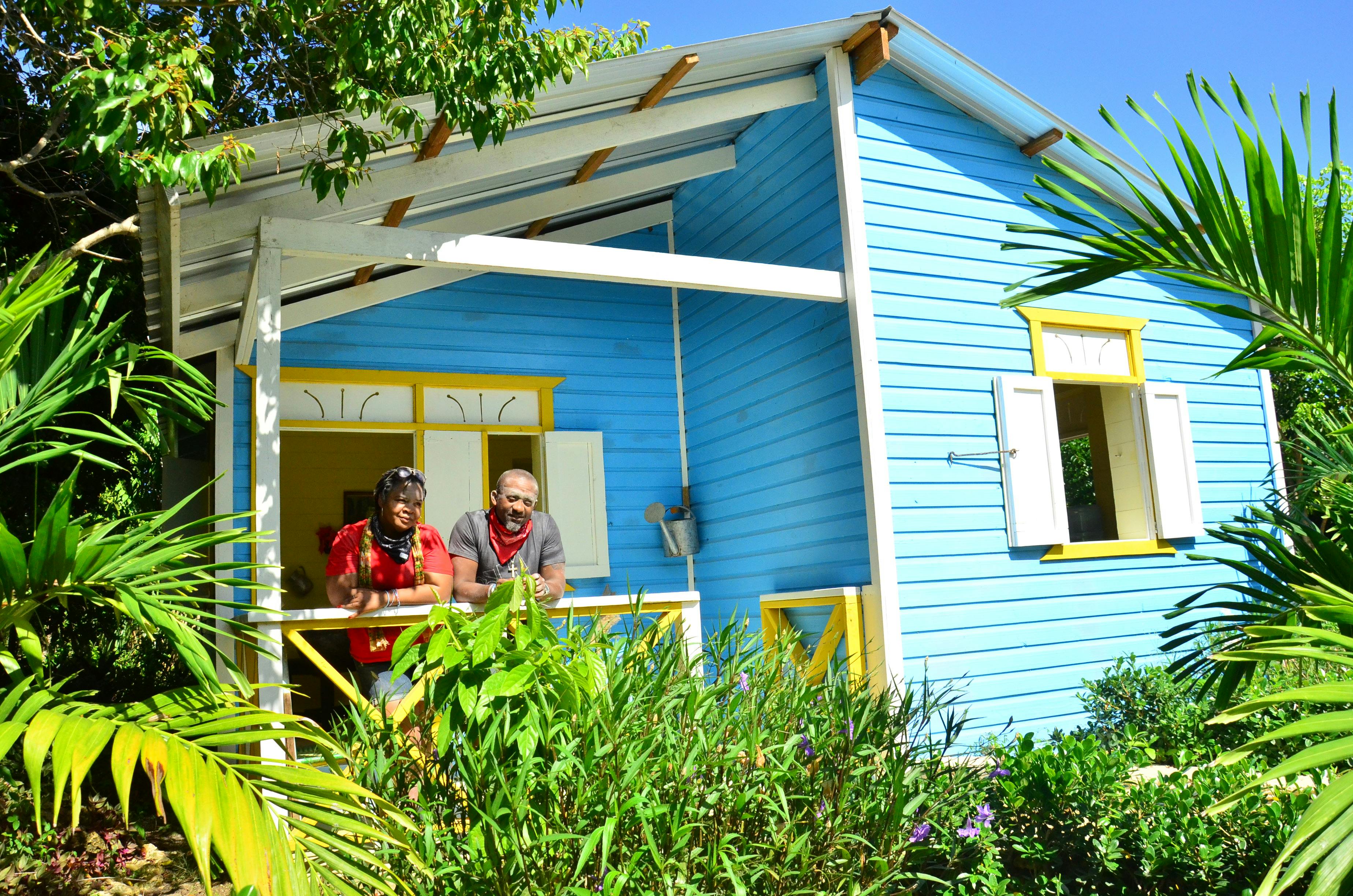 Two people on the porch of a vibrant blue and yellow house, surrounded by lush greenery under a clear blue sky.
