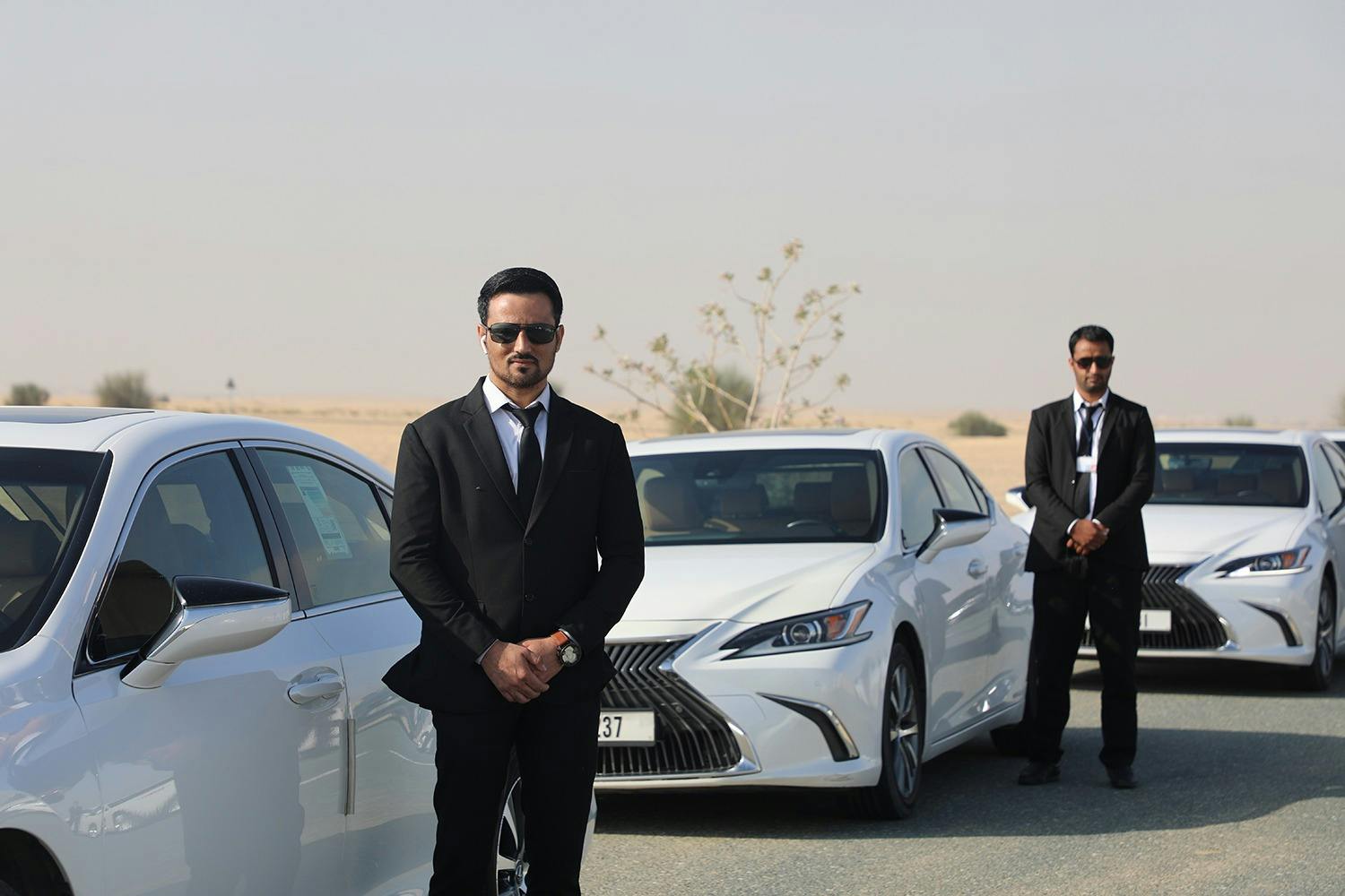 Man i svart kostym med solglasögon som står mellan två vita bilar i en ökenmiljö.