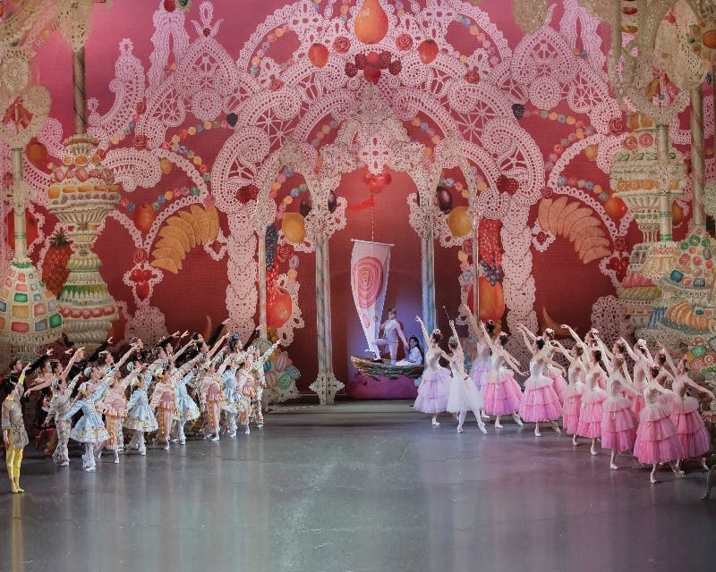 Ballet dancers dressed in colorful costumes perform on a stage with elaborate, pink, candy-themed decorations.