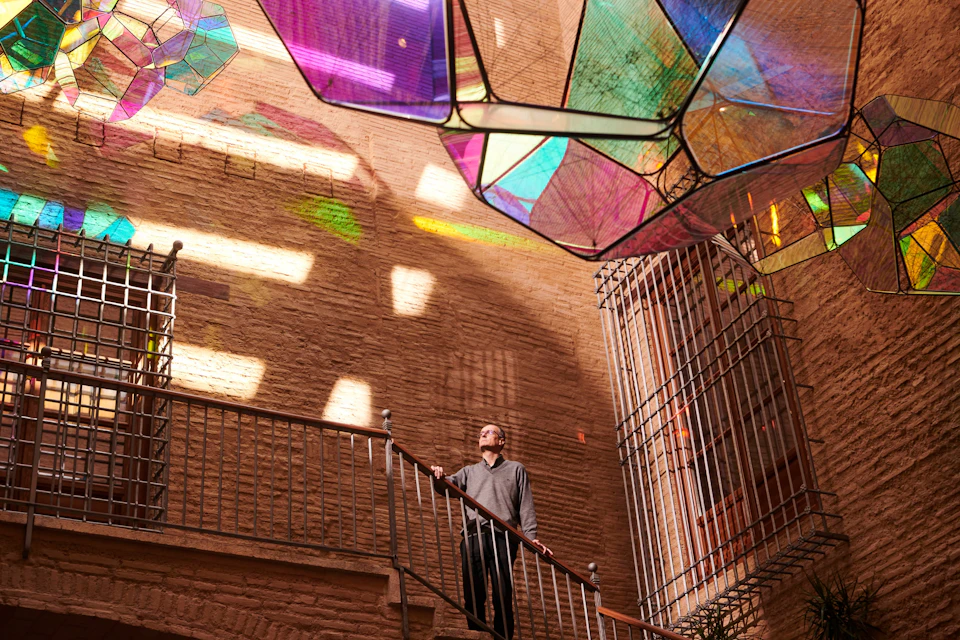A person stands on a staircase under colorful translucent geometric panels casting light patterns on brick walls.