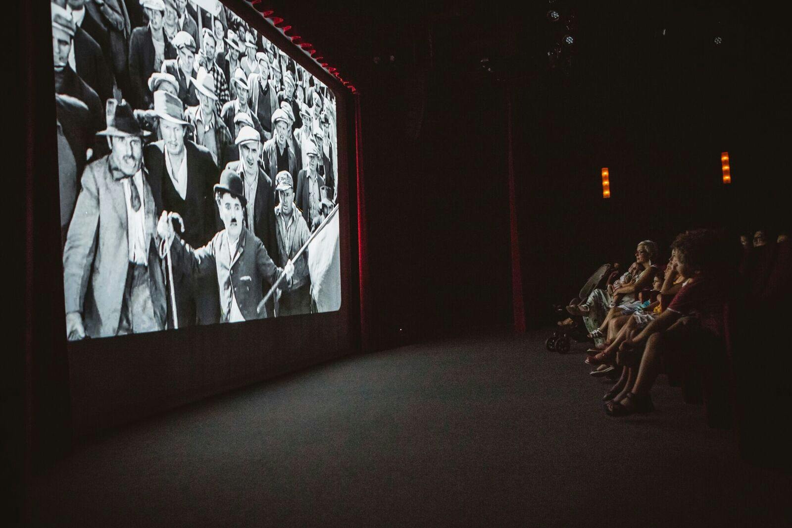 Lidé sledující černobílý film Charlieho Chaplina v temném kině. Publikum sedí po pravé straně.
