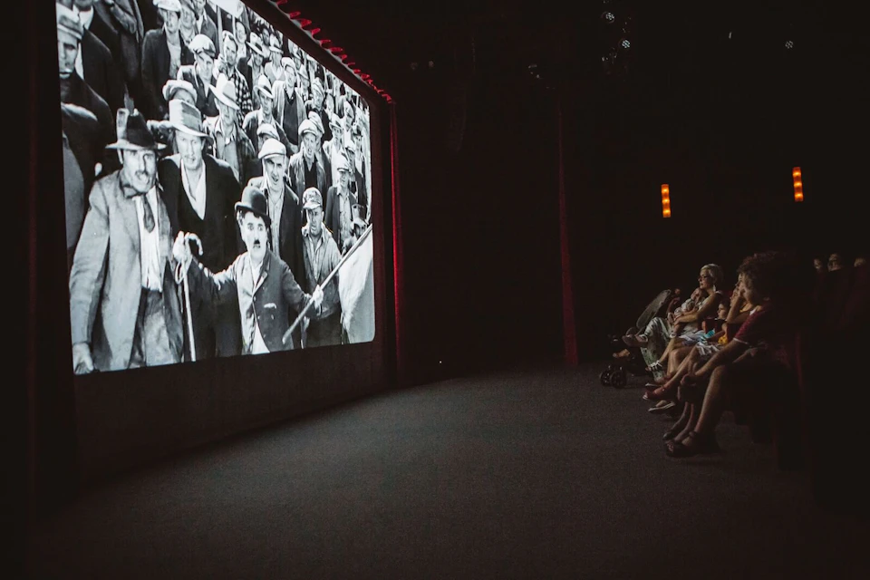 People watching a black-and-white Charlie Chaplin film in a dark theater. The audience is seated along the right side.