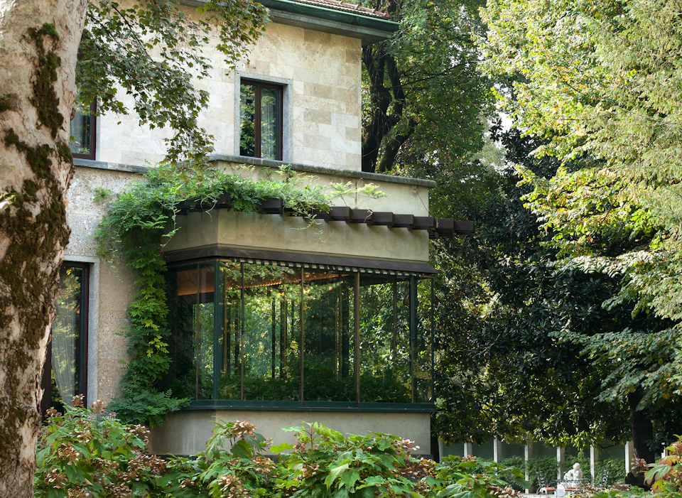 A stone building with a large, glass-enclosed sunroom surrounded by lush green plants and trees.