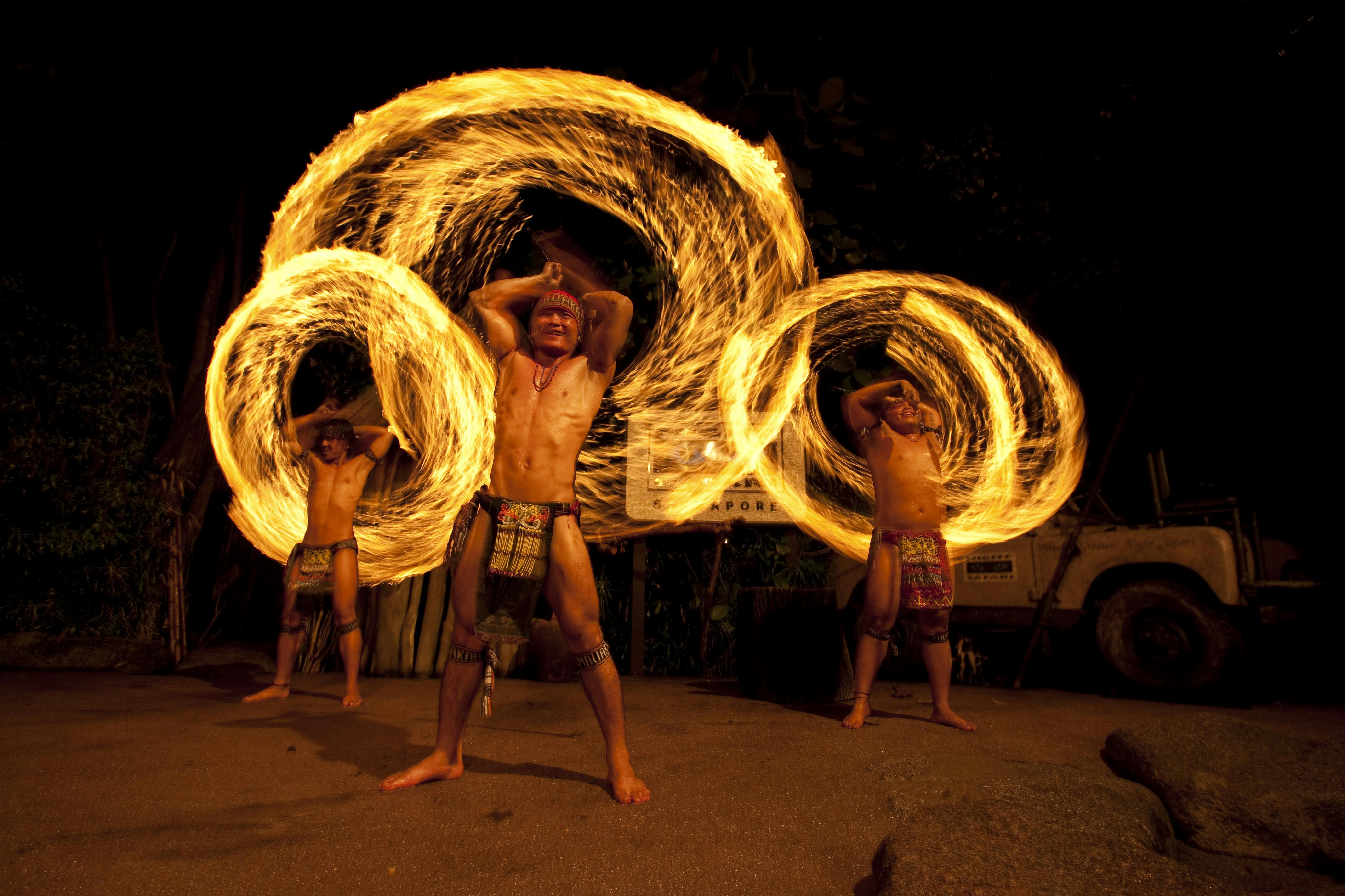 Dva umělci v tradičním oblečení točí ohnivé poi a v noci vytvářejí kruhové ohnivé stezky s vozidlem v pozadí.