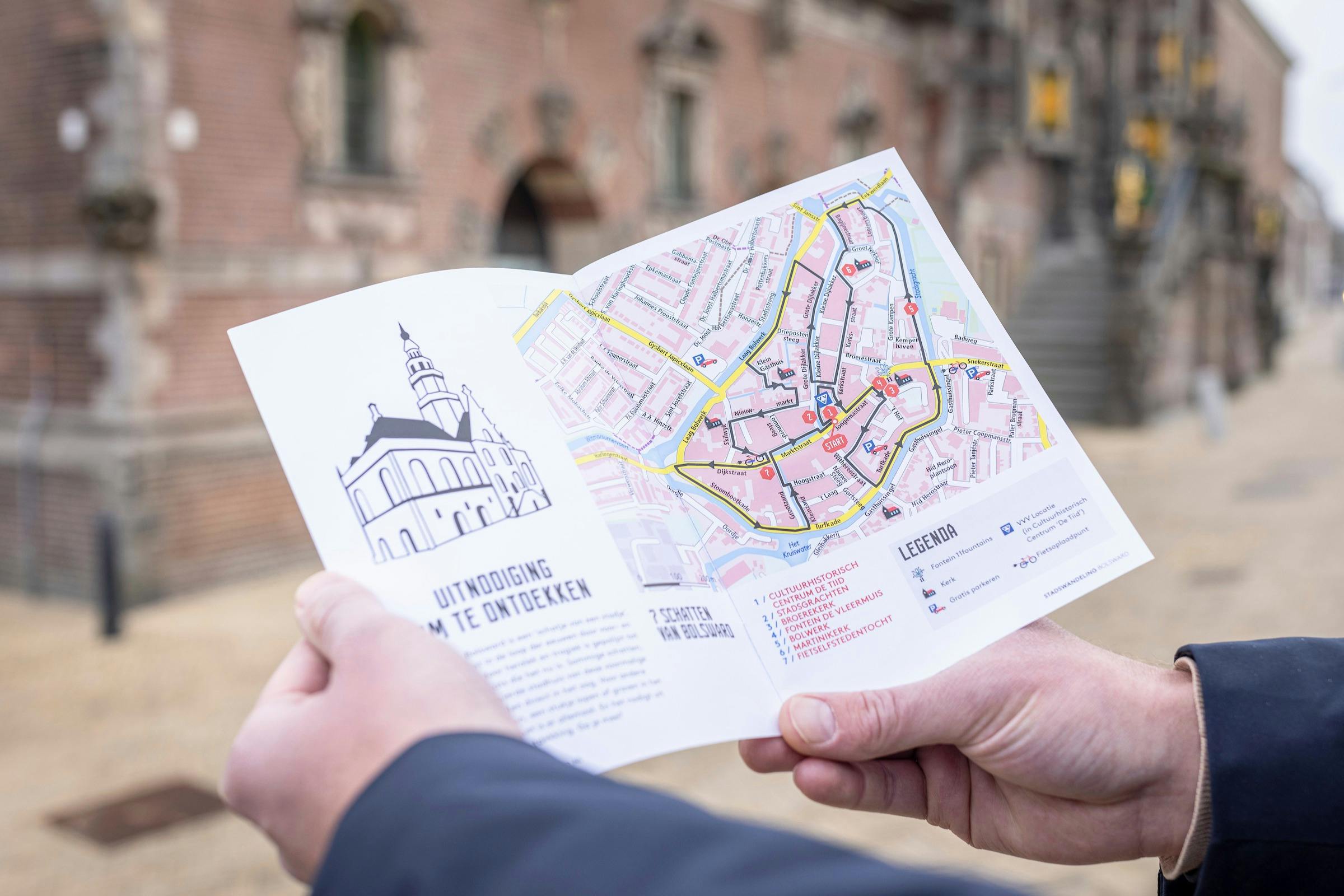Person holding an open map with a city layout, landmarks, and text in Dutch. Blurred building in the background.