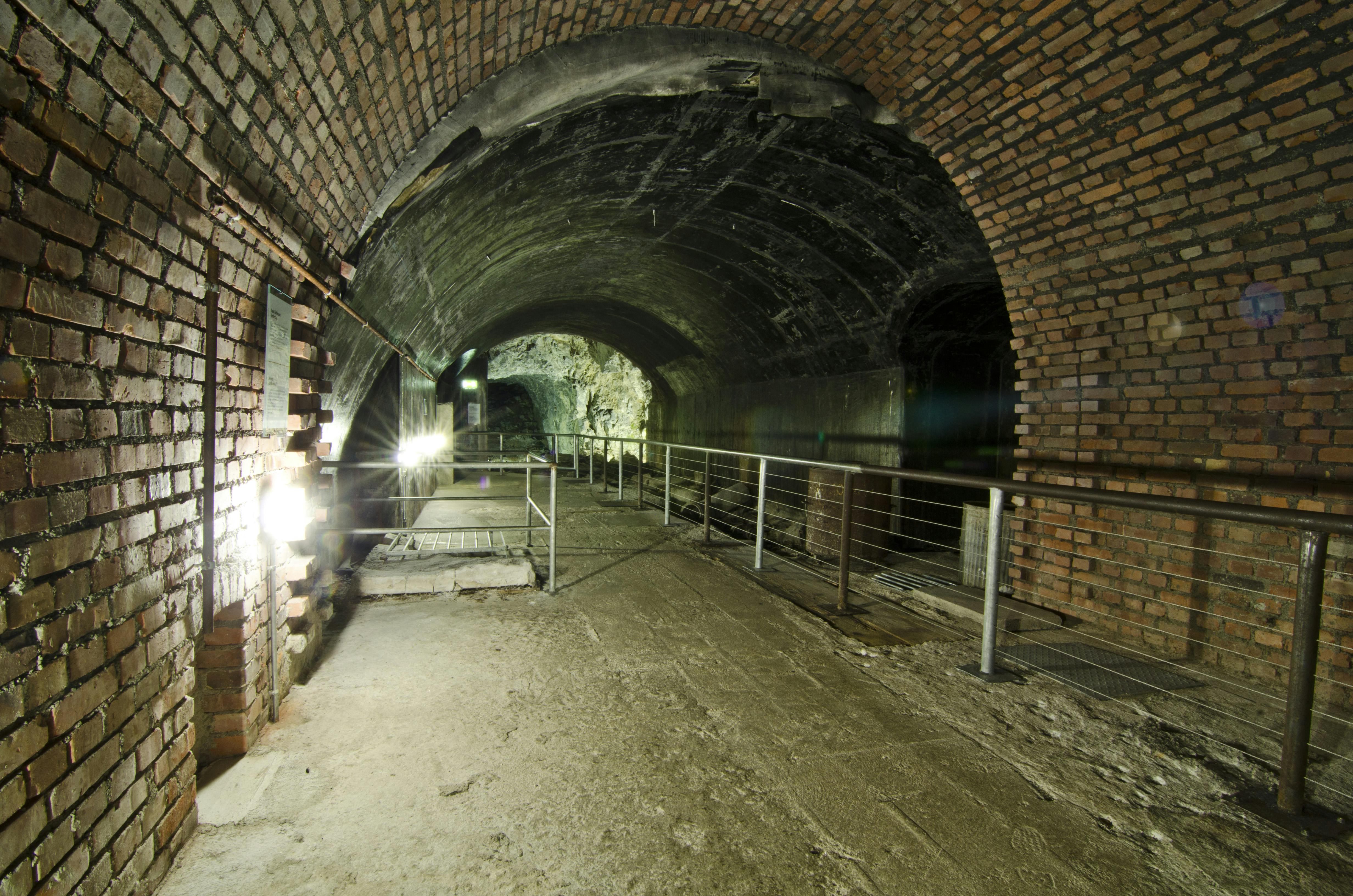 Brick-lined underground tunnel with arched ceiling, lit by overhead lights, and featuring a metal railing along a pathway.
