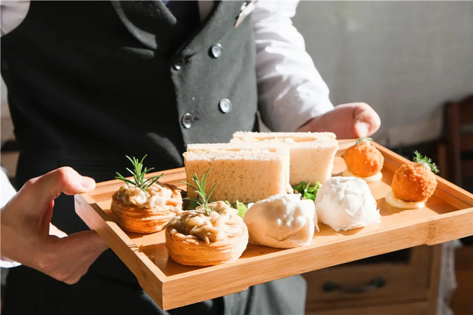 A server presenting a wooden tray with assorted pastries, sandwiches, and garnishes.