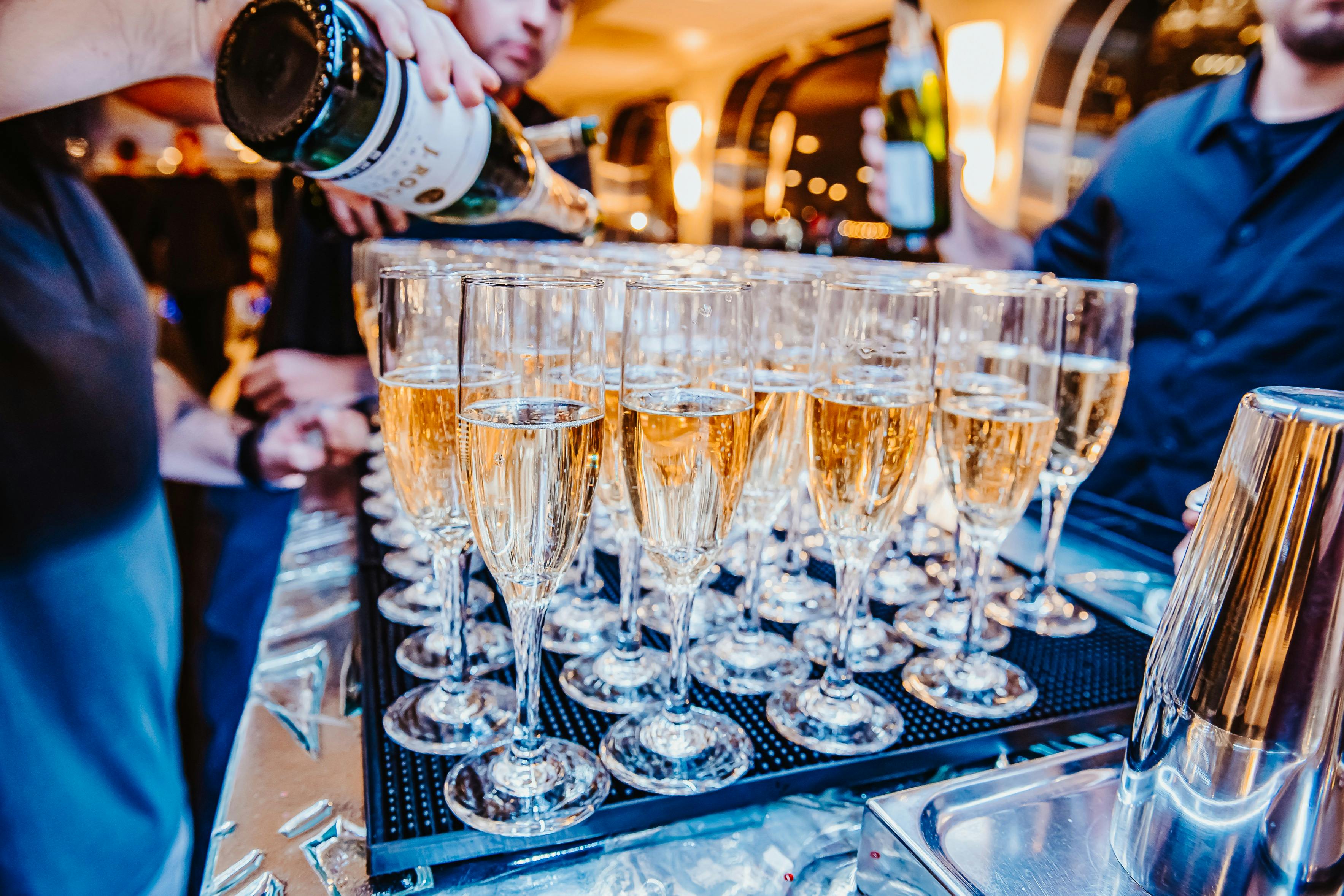 Několik sklenic šampaňského, které plní dva lidé v baru nebo na slavnostní akci, s hřejivým osvětlením v pozadí.