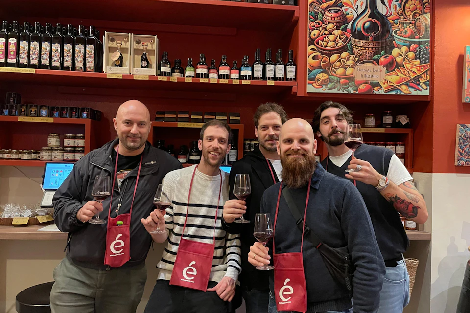 Five men smile and hold wine glasses in a wine shop, with shelves of bottles and colorful artwork behind them.