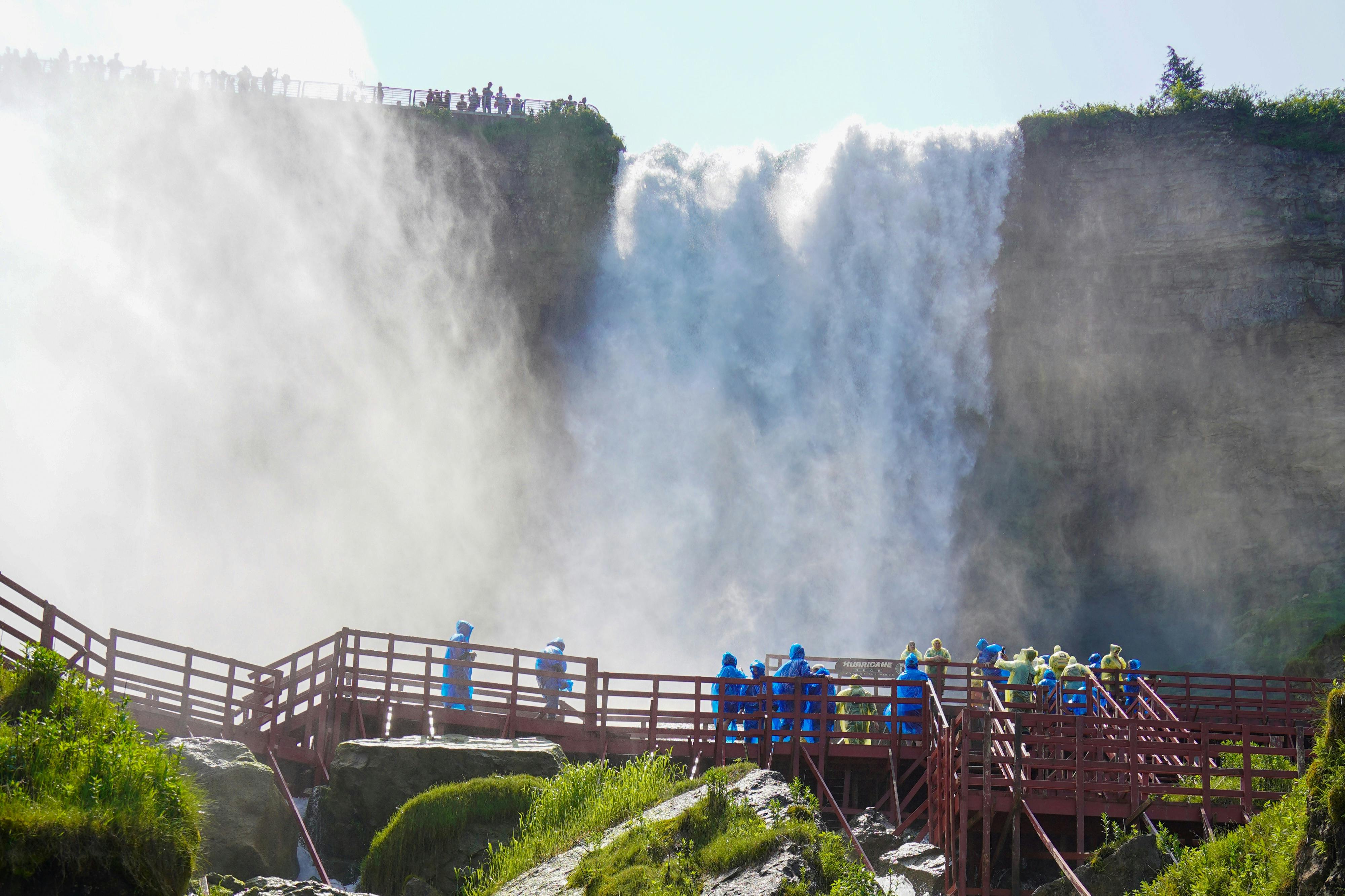 青と黄色のレインコートを着た人々は、大きな滝の近くの赤いボードウォークを歩いています。周りには霧が立ち上がっています。