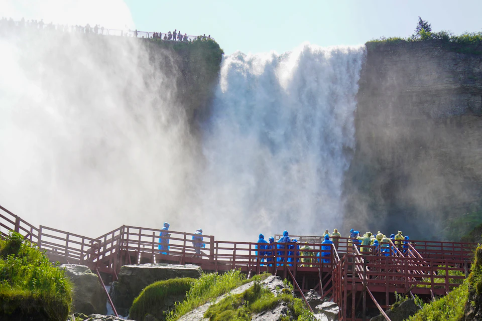 People in blue and yellow raincoats walk on a red boardwalk near a large waterfall, with mist rising around them.