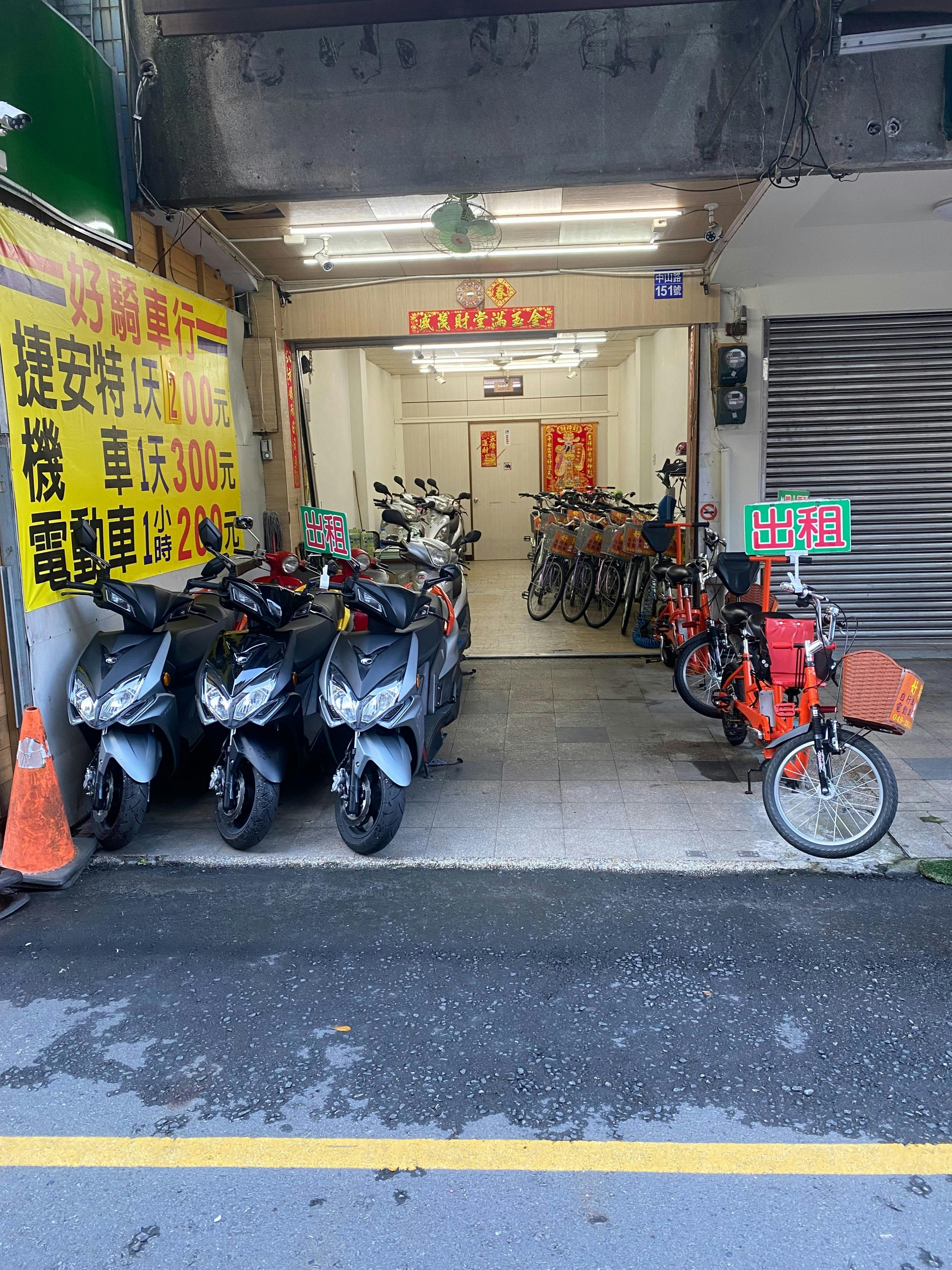 A storefront displaying rental bicycles and scooters, with a sign in Chinese and vehicles parked on the sidewalk.