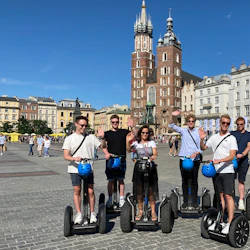 En grupp på sju personer på Segways vinkar på ett soligt torg med historiska byggnader och en kyrka i bakgrunden.