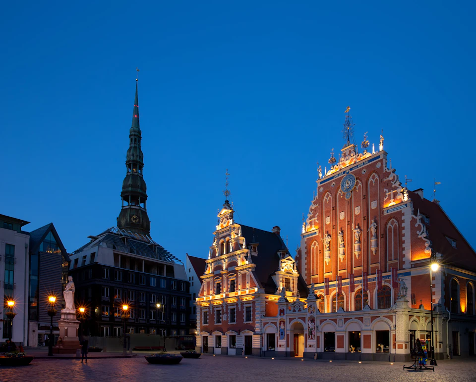 A historic square at dusk featuring illuminated Gothic buildings and a tall, slender spire against a deep blue sky.