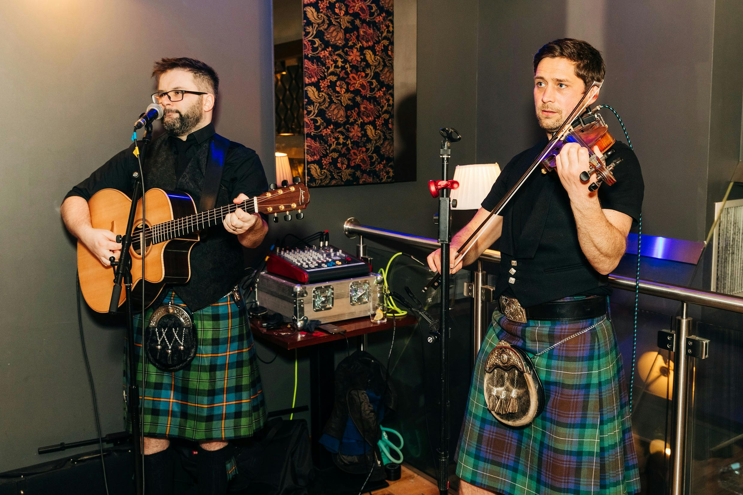 Un homme en kilt joue du violon, debout à l'intérieur, avec du matériel de sonorisation visible derrière lui.