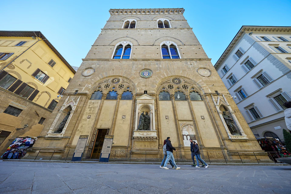 A historic building with statues in niches and arched windows, viewed from below, with people walking in the foreground.