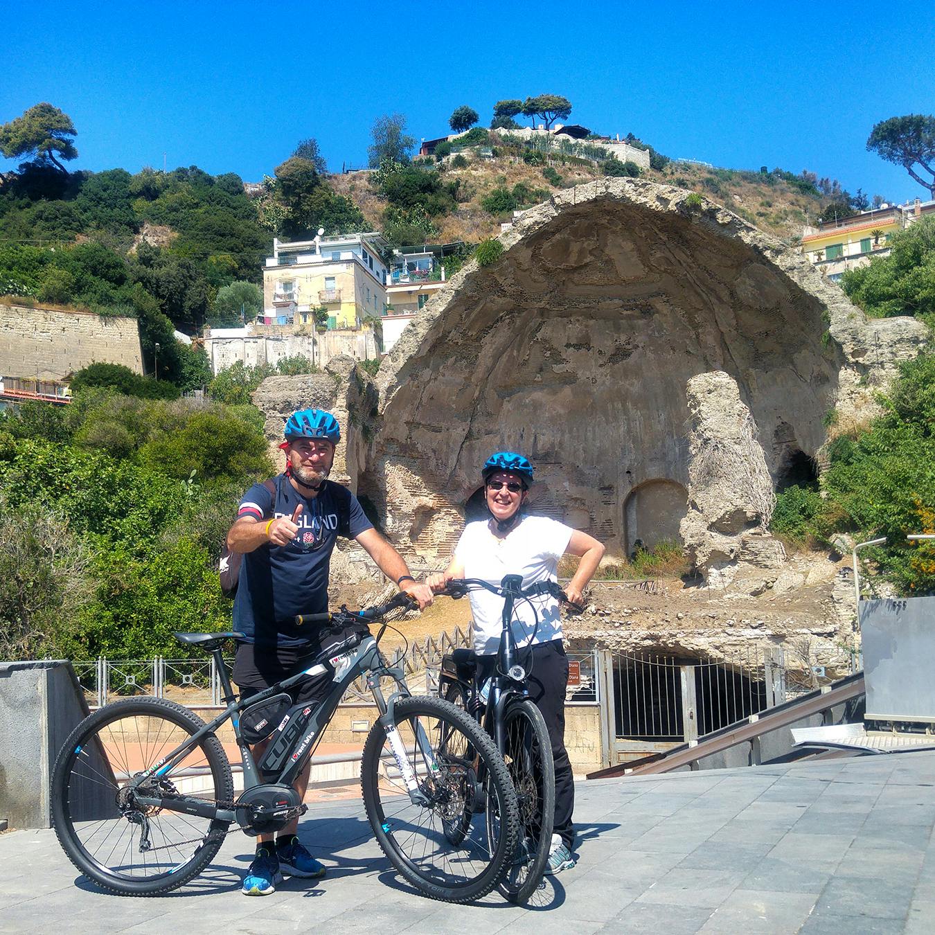 Due persone in tenuta da ciclismo e casco si trovano con le loro biciclette su un sentiero asfaltato, sorridendo, con una collina rocciosa e rovine di edifici alle loro spalle.