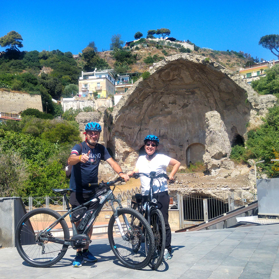 Two people in cycling gear and helmets stand with their bikes on a paved path, smiling, with a rocky hill and building ruins behind them.