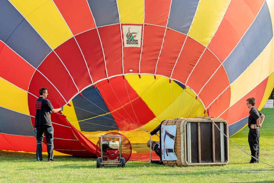 A person prepares a colorful hot air balloon for inflation on a grassy field; a basket and fan are in the foreground.
