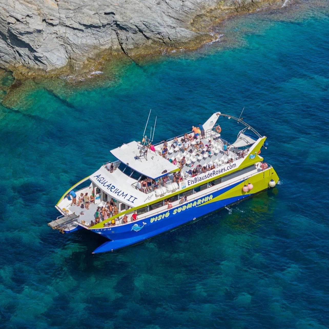 A large yellow and blue boat named Aquarium II carrying passengers over clear blue water near a rocky shore.