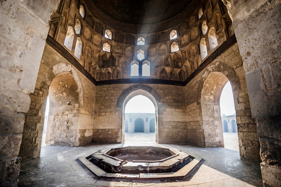 Historic stone building interior with sunlight streaming through intricate windows, arched doorways, and a central octagonal basin.