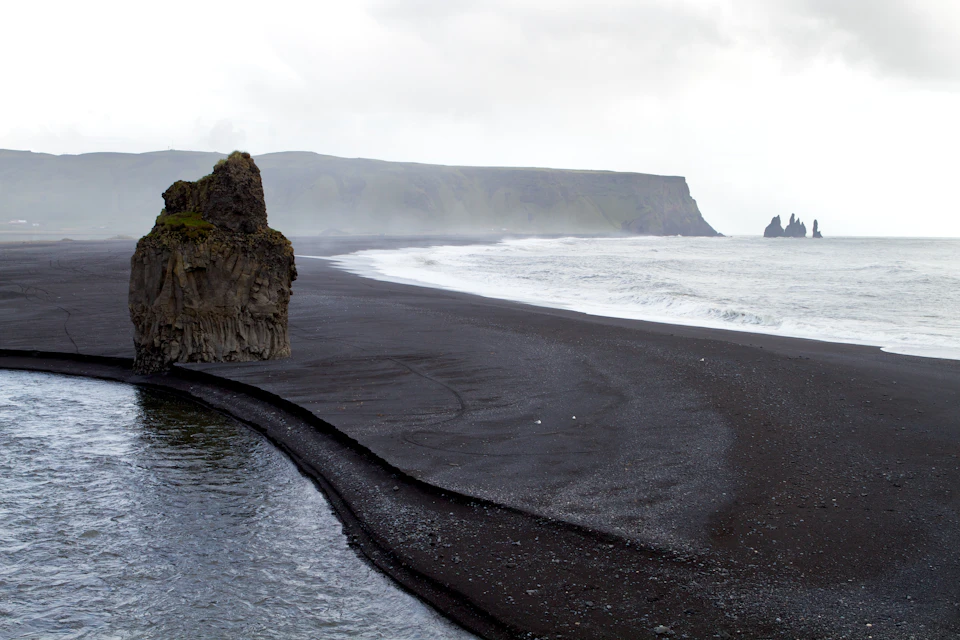 Black sand beach with rugged cliffs, distant sea stacks, and overcast sky. Waves gently lap the shore.
