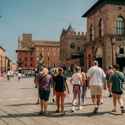 People walking in a square with historic buildings, and a statue in the background, on a sunny day.