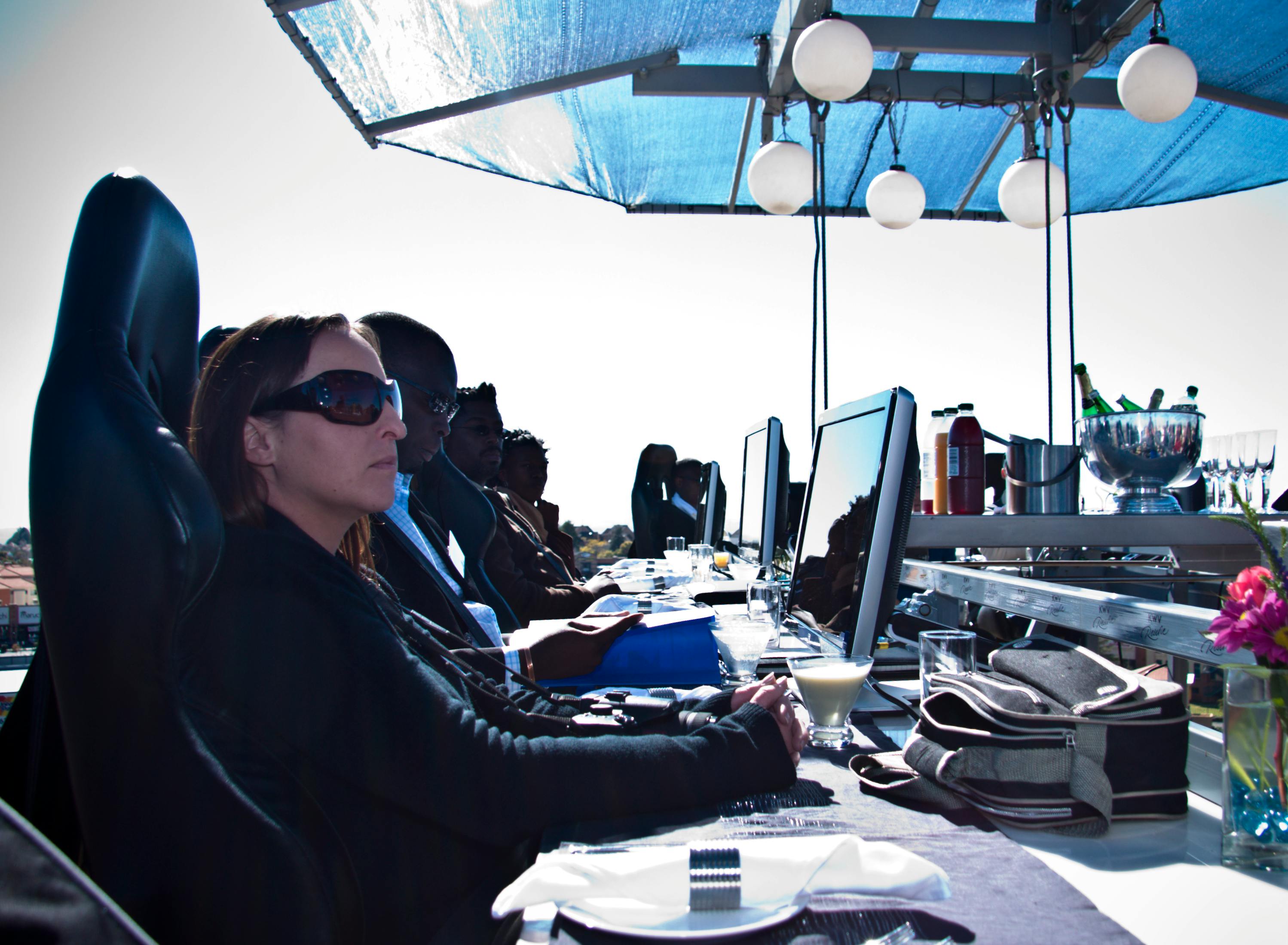 Människor som sitter vid ett högt bord med datorer och drycker, under ett tak med hängande lampor, vänd åt samma håll utomhus.