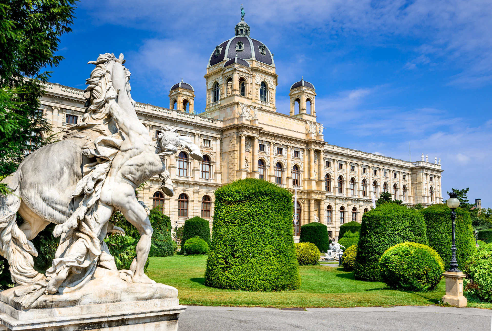 Historic building with large dome and statues, surrounded by manicured hedges and statues, under a bright blue sky.