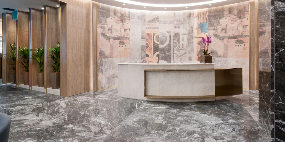 A sleek lobby with a curved marble reception desk, ornate wall art, polished marble floors, and a potted plant with purple flowers.