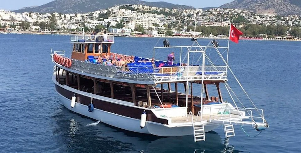 People relax on a docked boat with a multi-level deck in a coastal area with buildings and mountains in the background.
