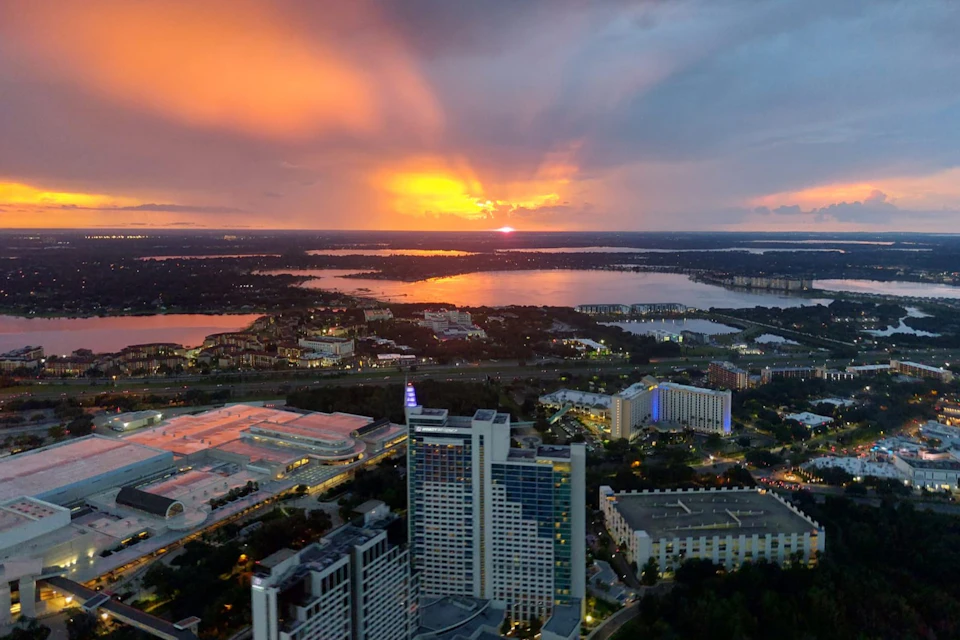 Sunset over a cityscape with high-rise buildings and a large body of water in the distance beneath a colorful sky.