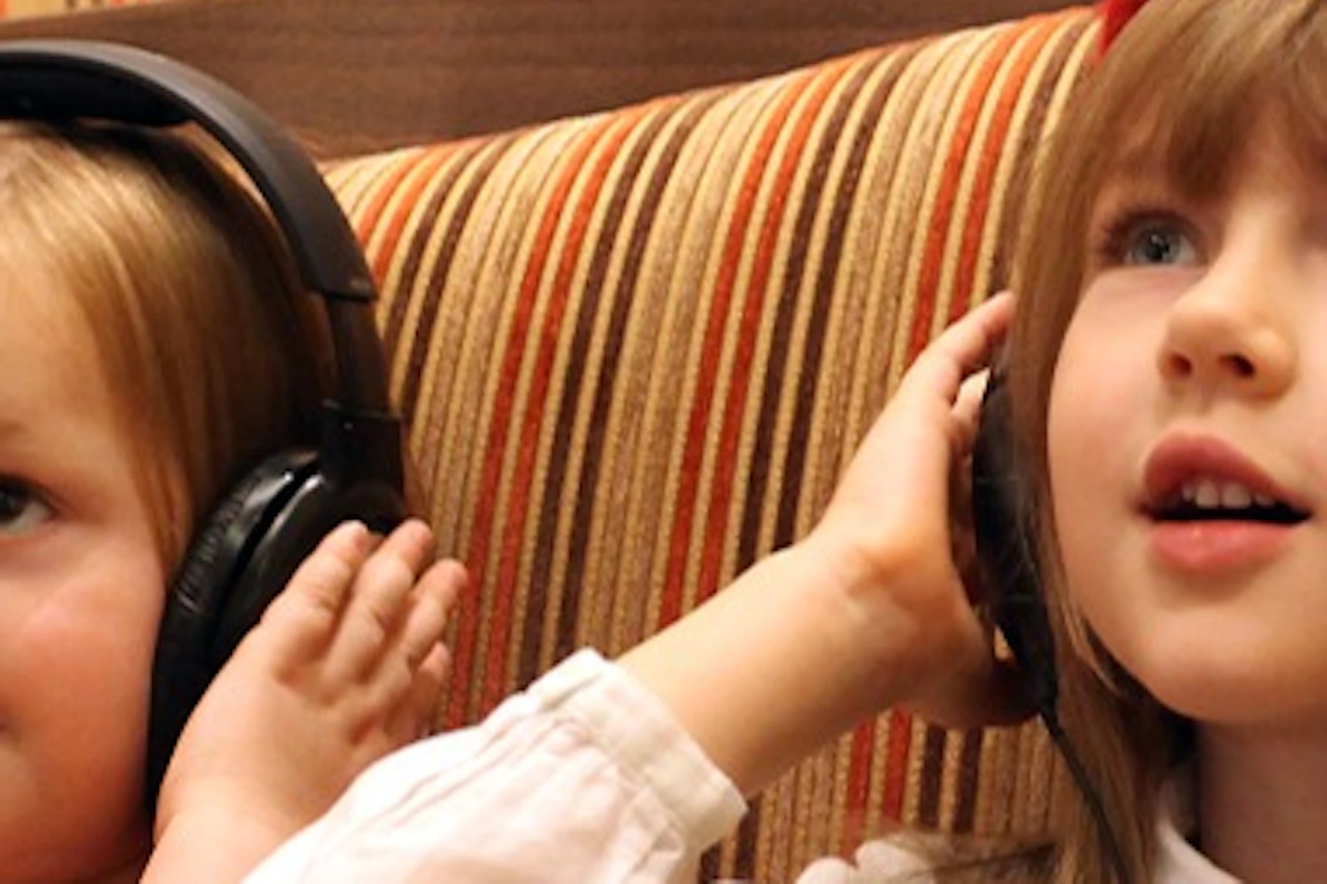 Two young girls sitting on a striped seat, wearing headphones, with one wearing a red beret and looking attentive.