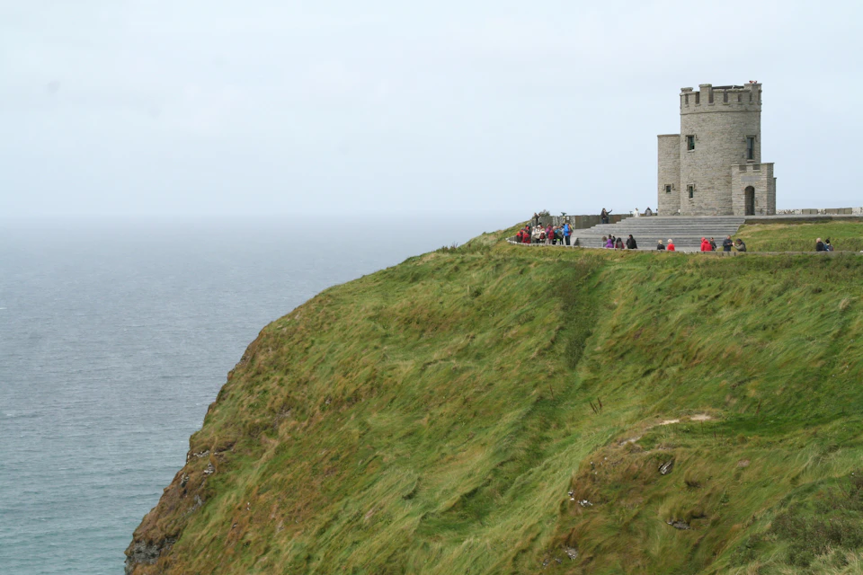 Green cliffside with stone building, tourists gathered near the edge overlooking the ocean.