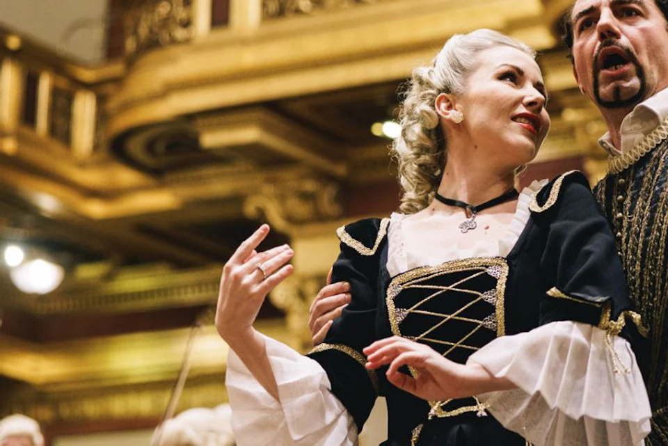 Two performers in ornate period costumes sing passionately on stage, with a classical orchestra and gilded decor in the background.