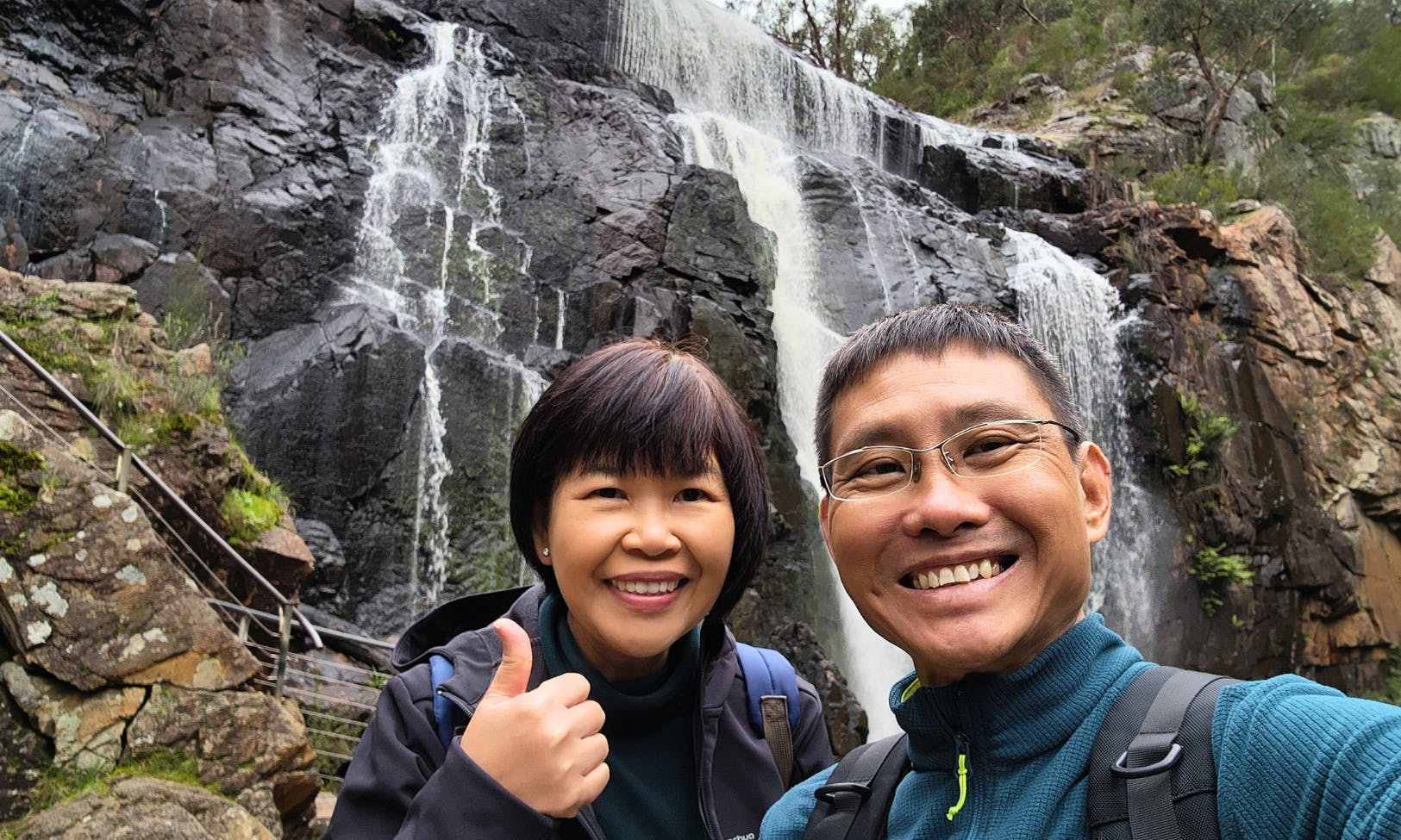 两个微笑的人在瀑布前摆姿势，背景是岩石地形和绿色植物。