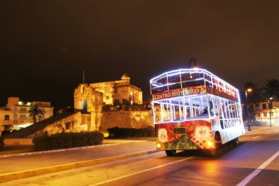 A double-decker tour bus with bright lights labeled "Centro Historico" is parked near an illuminated historic fortress at night.