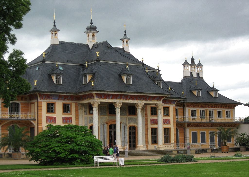 Ornate building with dark slate roofs, decorated columns, and a garden in front. Several people stand near white benches.