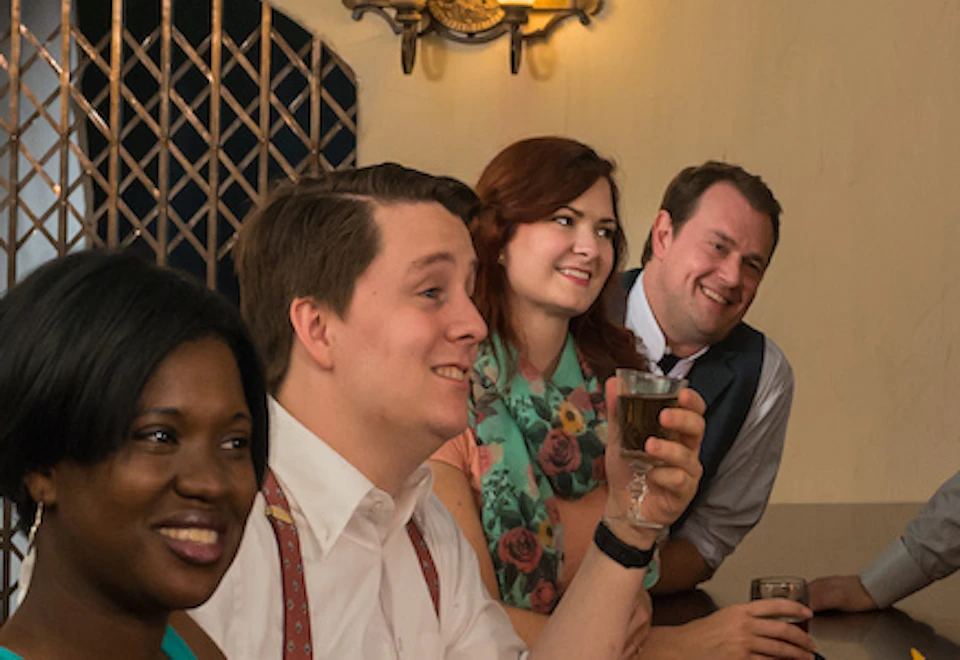 A group of people sits at a bar, holding drinks and smiling, with a bartender serving them in a cozy, dimly lit setting.