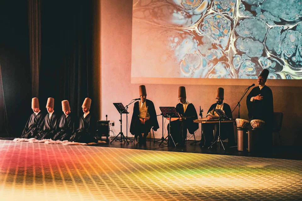 Musicians in traditional attire perform on a stage with instruments under a colorful abstract projection.