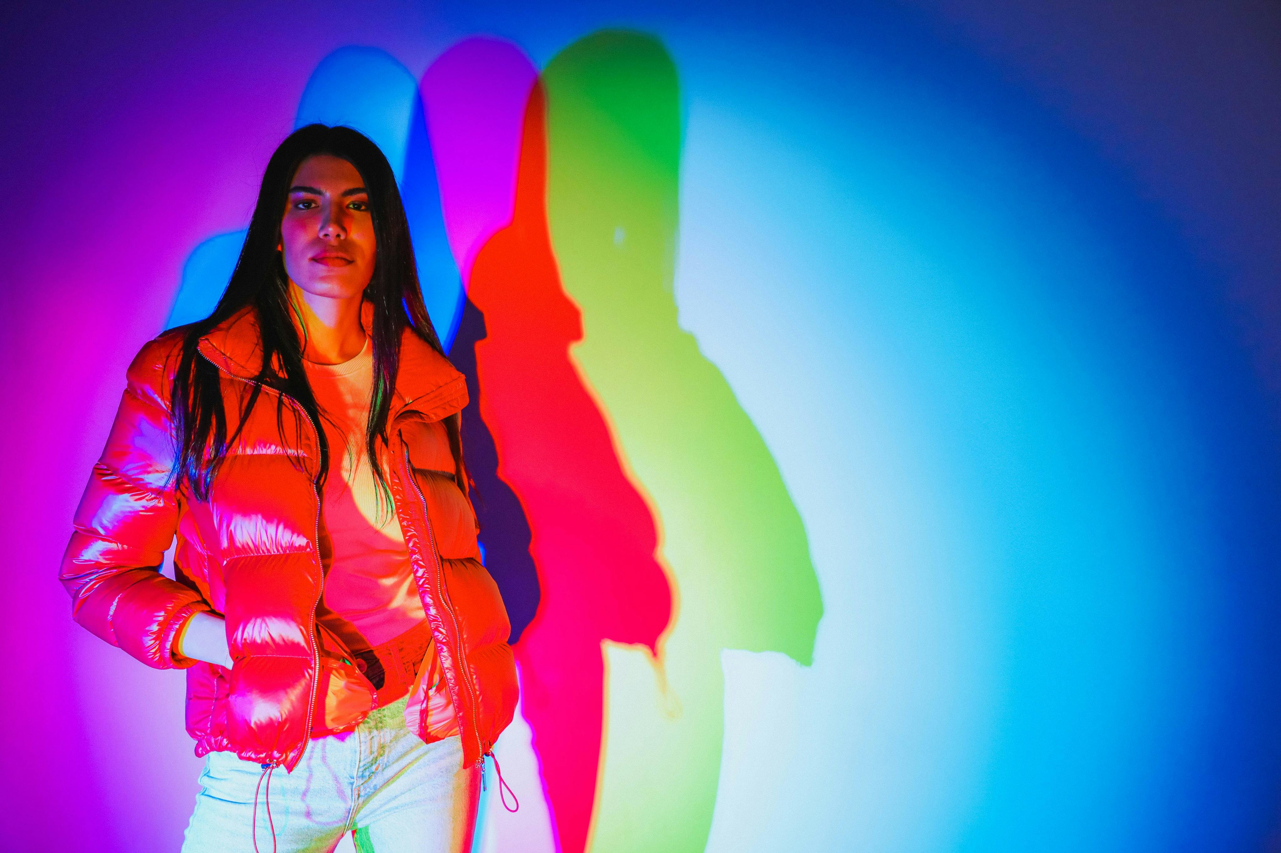 Una persona con una chaqueta acolchada roja está de pie junto a una pared, con luces de colores que crean sombras superpuestas en rojo, azul y verde.