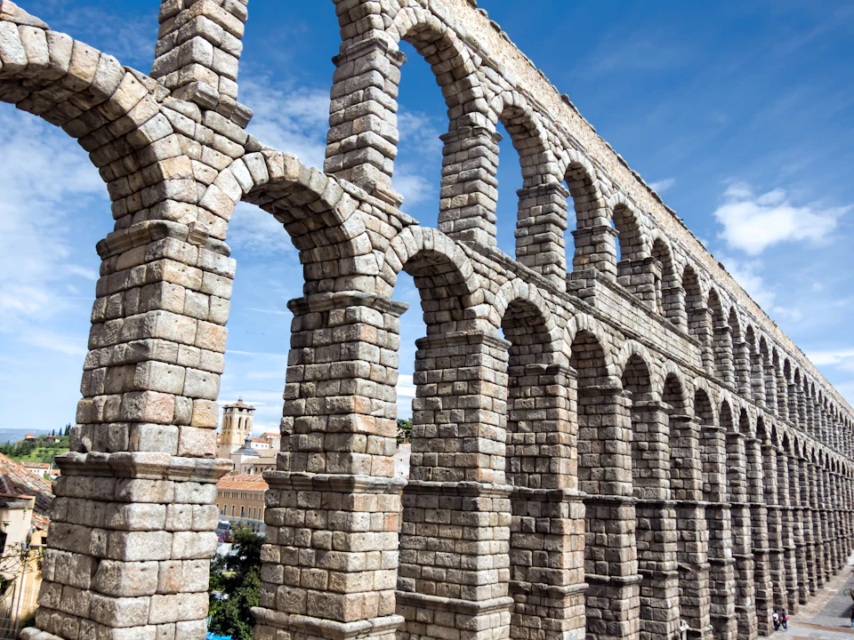 Stone aqueduct with multiple arches set against a blue sky. Buildings are visible in the background.
