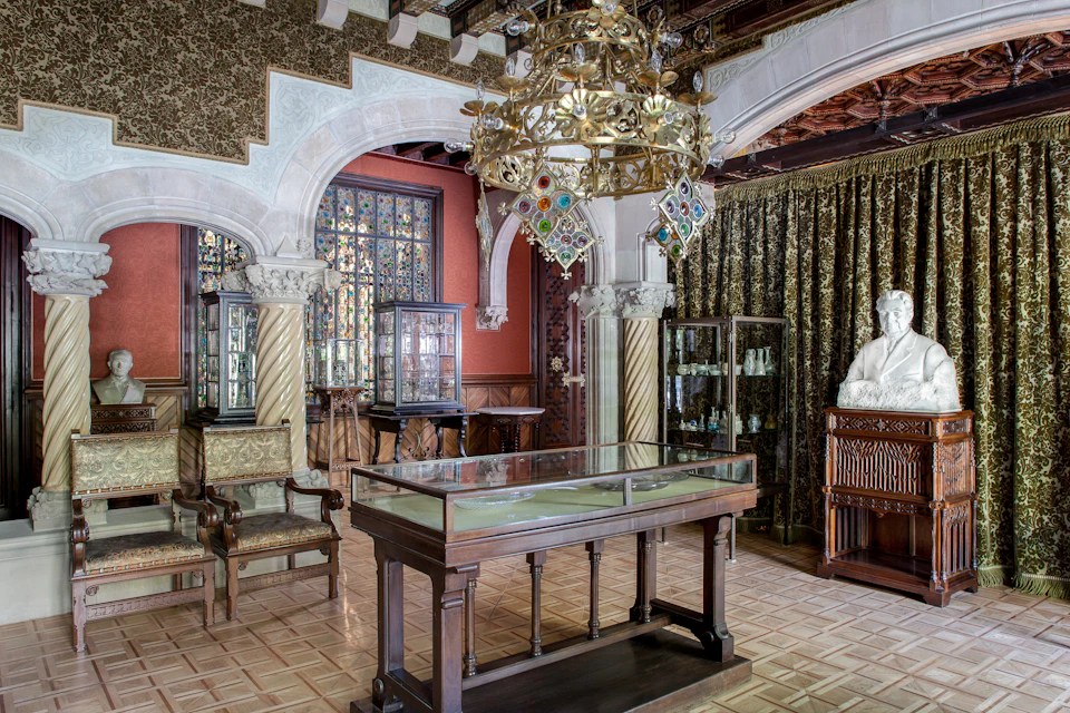 Opulent room with ornate chandeliers, display cases with glass items, carved chairs, marble columns, and a white bust on a wooden pedestal.