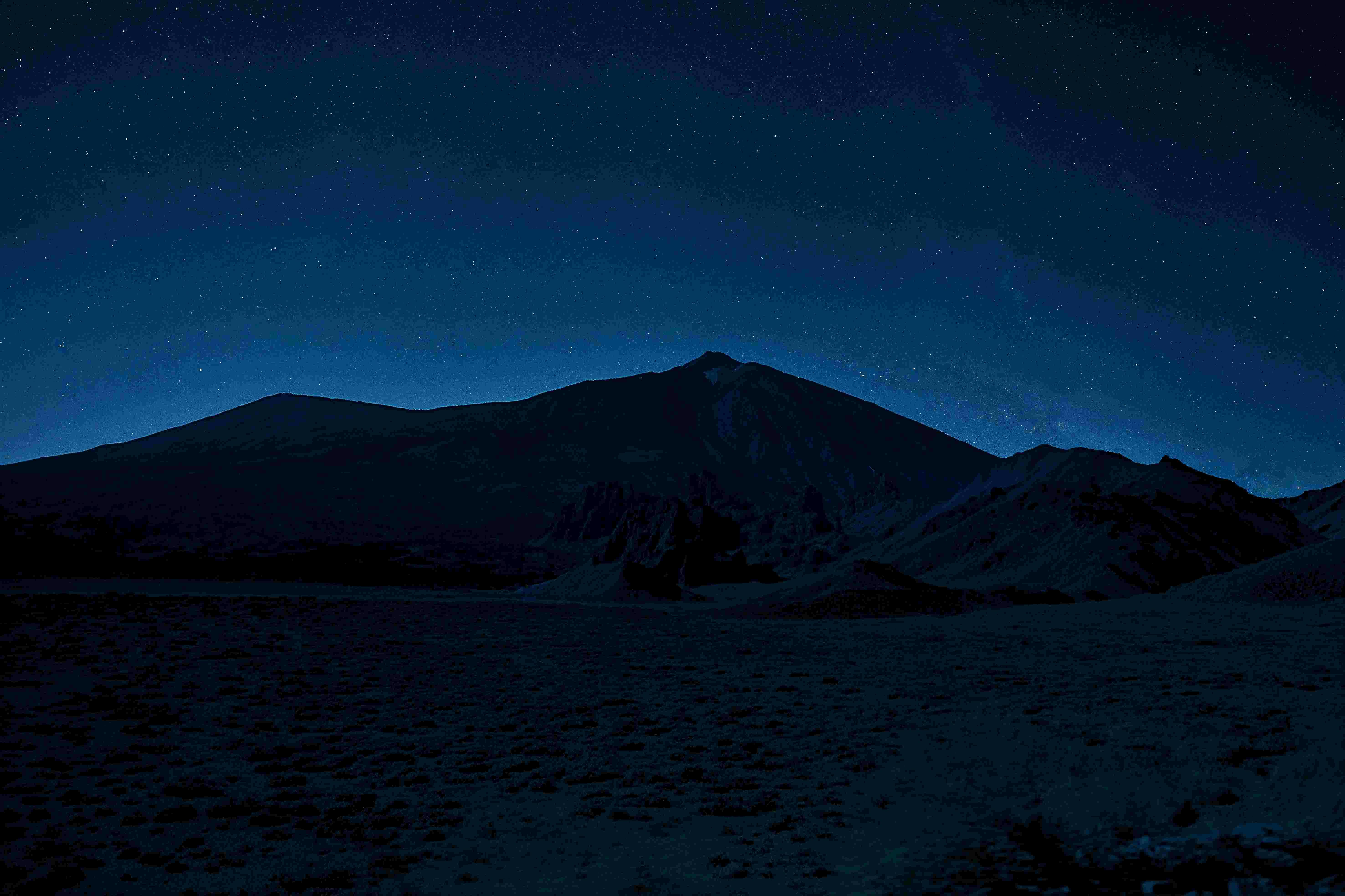 A mountain range silhouetted against a starry night sky, with a landscape of rocks and sparse vegetation in the foreground.